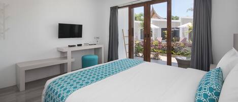 A modern hotel room with a large bed, a desk, a television, and a sliding glass door leading to a balcony with plants.