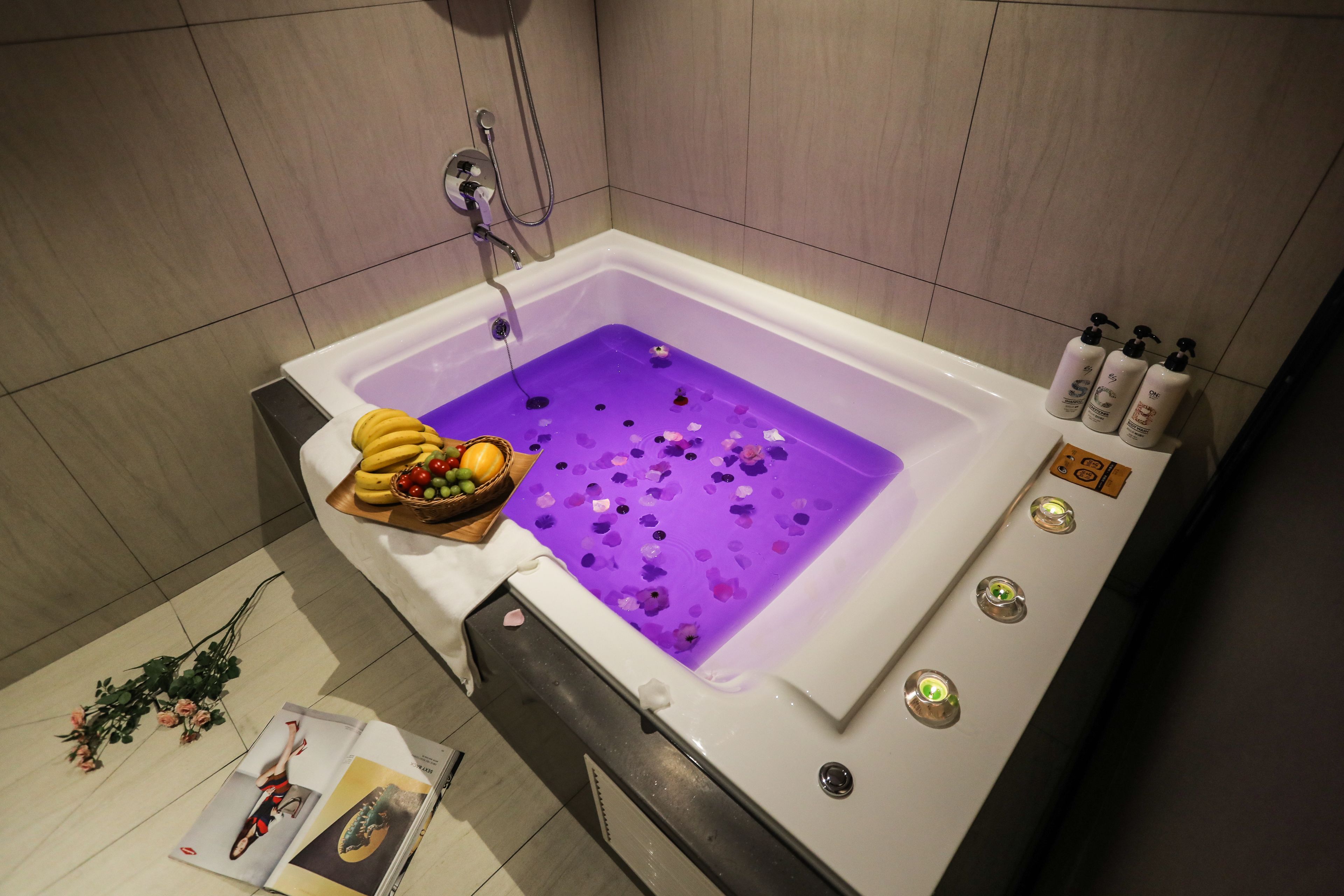 A modern bathroom with a bathtub filled with purple bath salts, a fruit bowl, and a magazine on the floor.