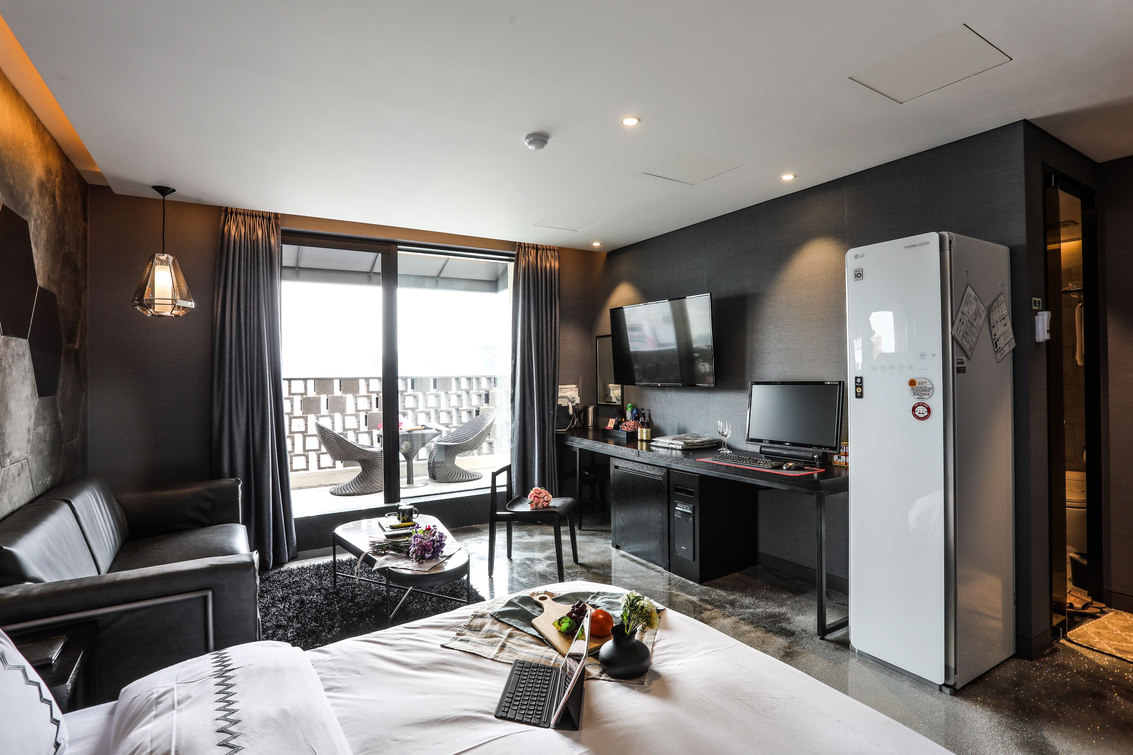 A modern hotel room with a bed, a desk, a refrigerator, a TV, and a view of the city.