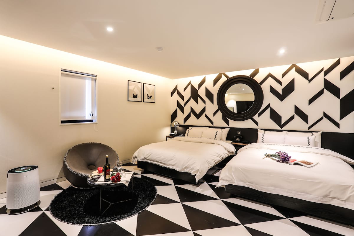 A modern bedroom with a black and white chevron-patterned wall, a round mirror, a bed with white bedding, and a black round table with a bottle and flowers.