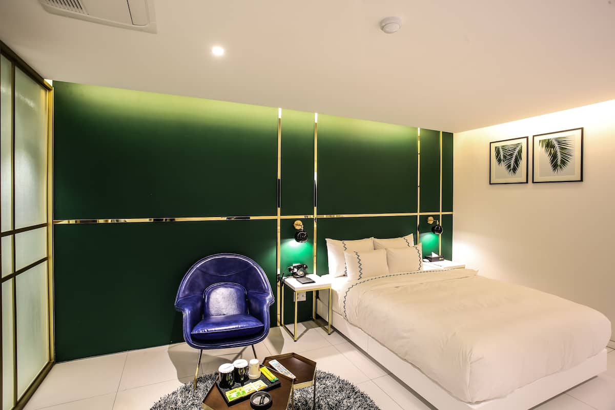 A modern hotel room with a green accent wall, a white bed, a blue chair, a bedside table with a lamp, and two framed pictures on the wall.