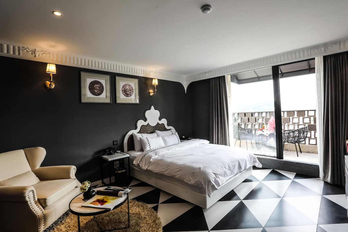 A hotel room with a bed, a sofa, a small table with a vase and book, and a balcony with outdoor seating.