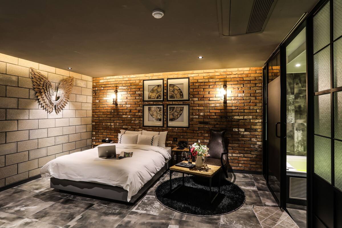 A modern bedroom with a brick wall, a large bed, a desk, and a view through a sliding door.