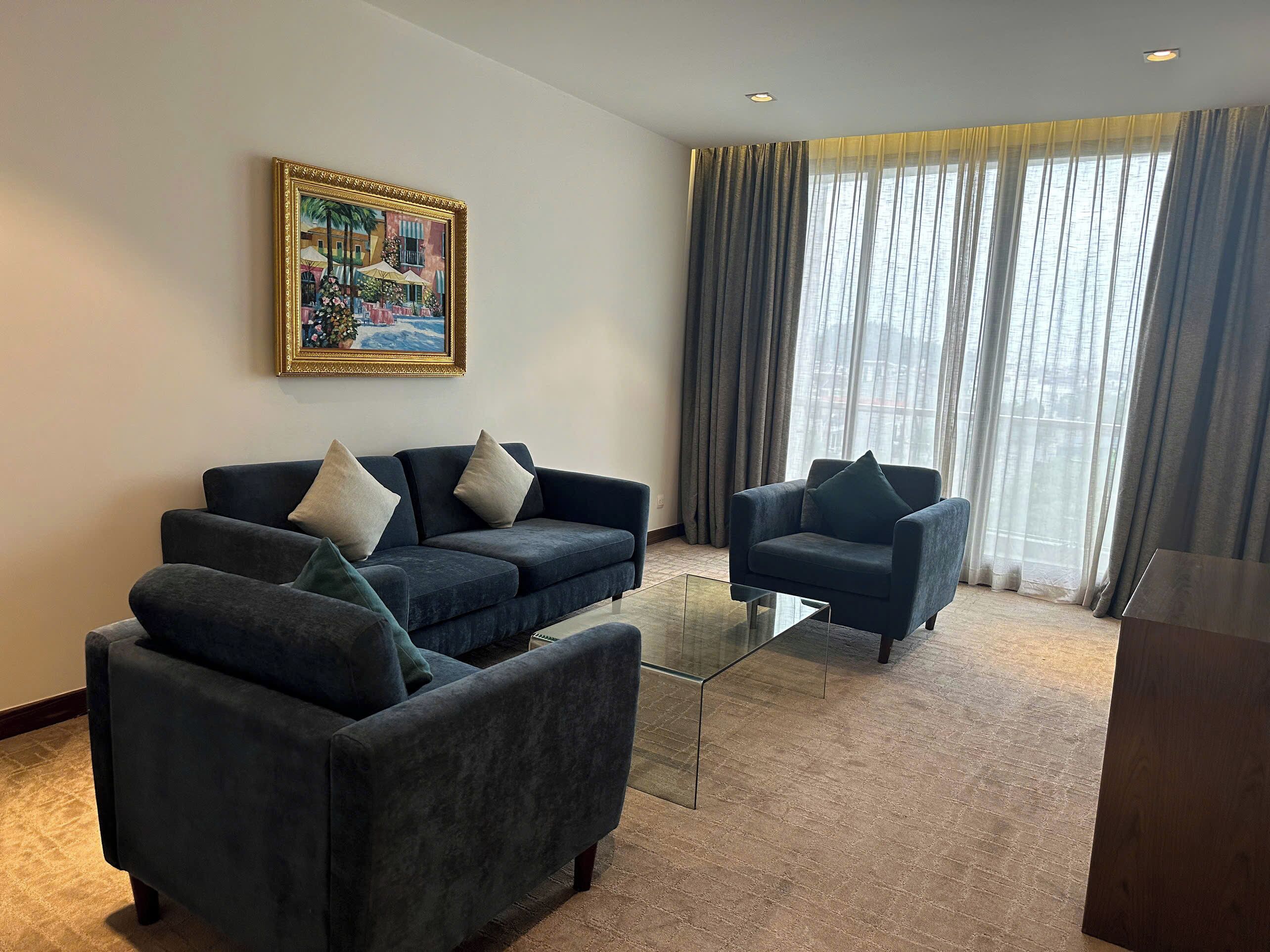 A modern living room with a sofa and armchair, a glass coffee table, a painting on the wall, and a large window with curtains.