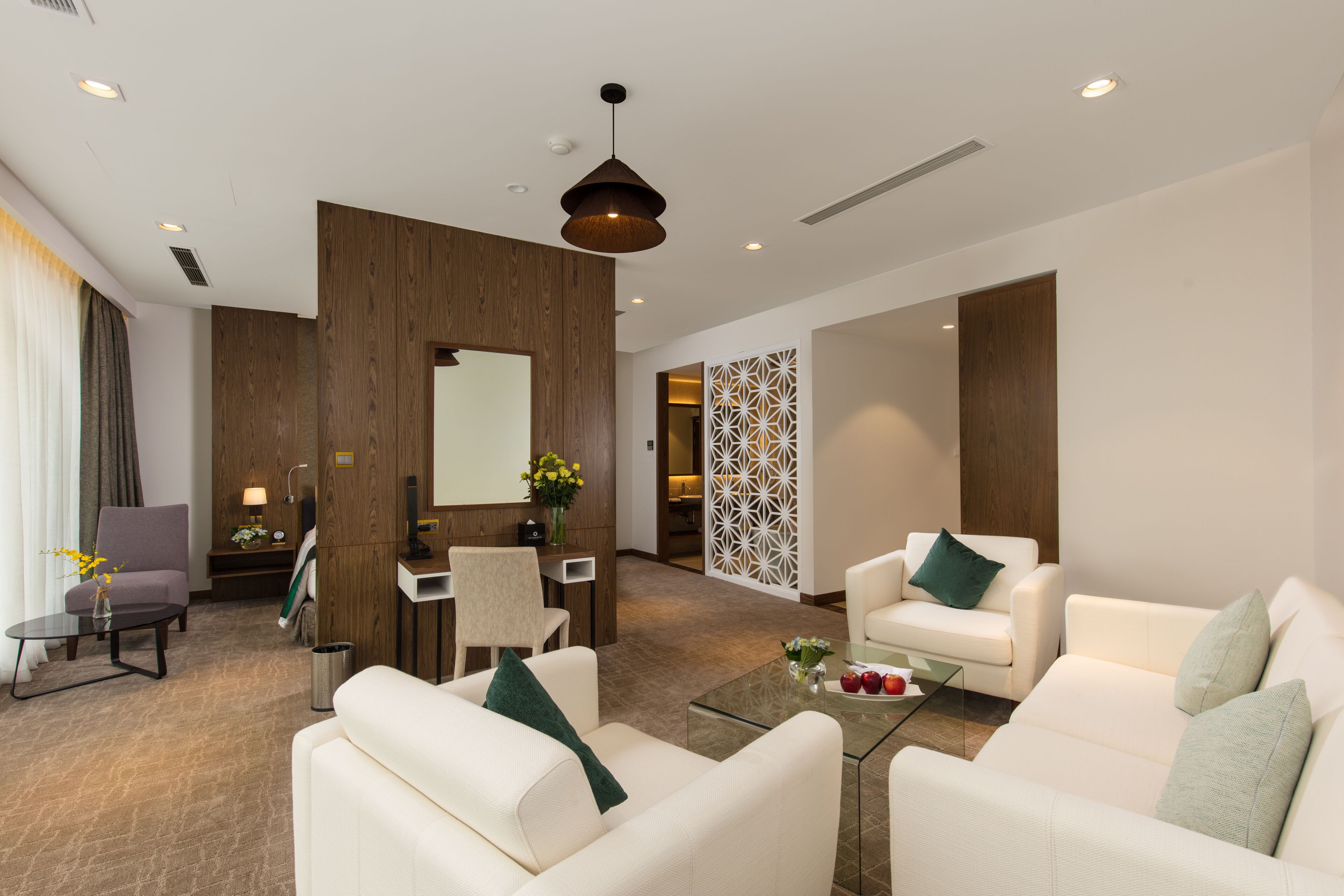 A modern hotel lobby with a reception desk, two white sofas with green cushions, a coffee table, and a decorative wall panel.