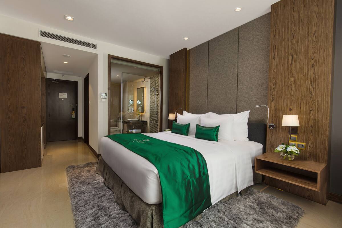 A modern hotel room with a large bed, a wooden headboard, a nightstand with a lamp, and a mirror.