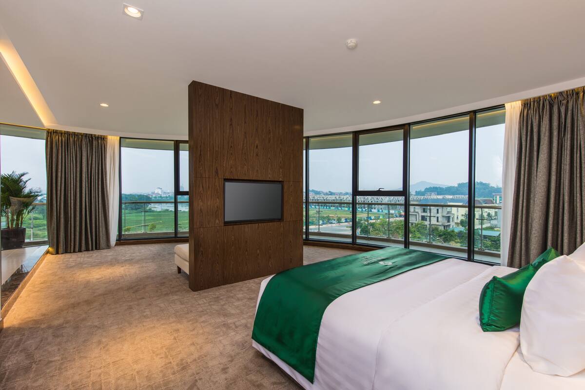 A modern hotel room with a large bed, a flat-screen TV mounted on a wooden panel, and a view of a cityscape through large windows.