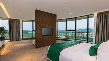 A modern hotel room with a large bed, a flat-screen TV mounted on a wooden panel, and a view of a cityscape through large windows.
