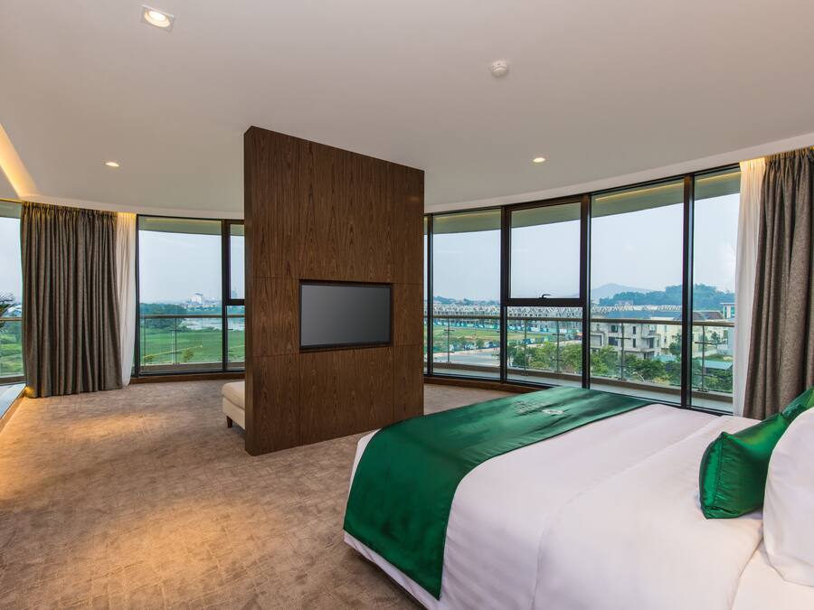 A modern hotel room with a large bed, a flat-screen TV mounted on a wooden panel, and a view of a cityscape through large windows.