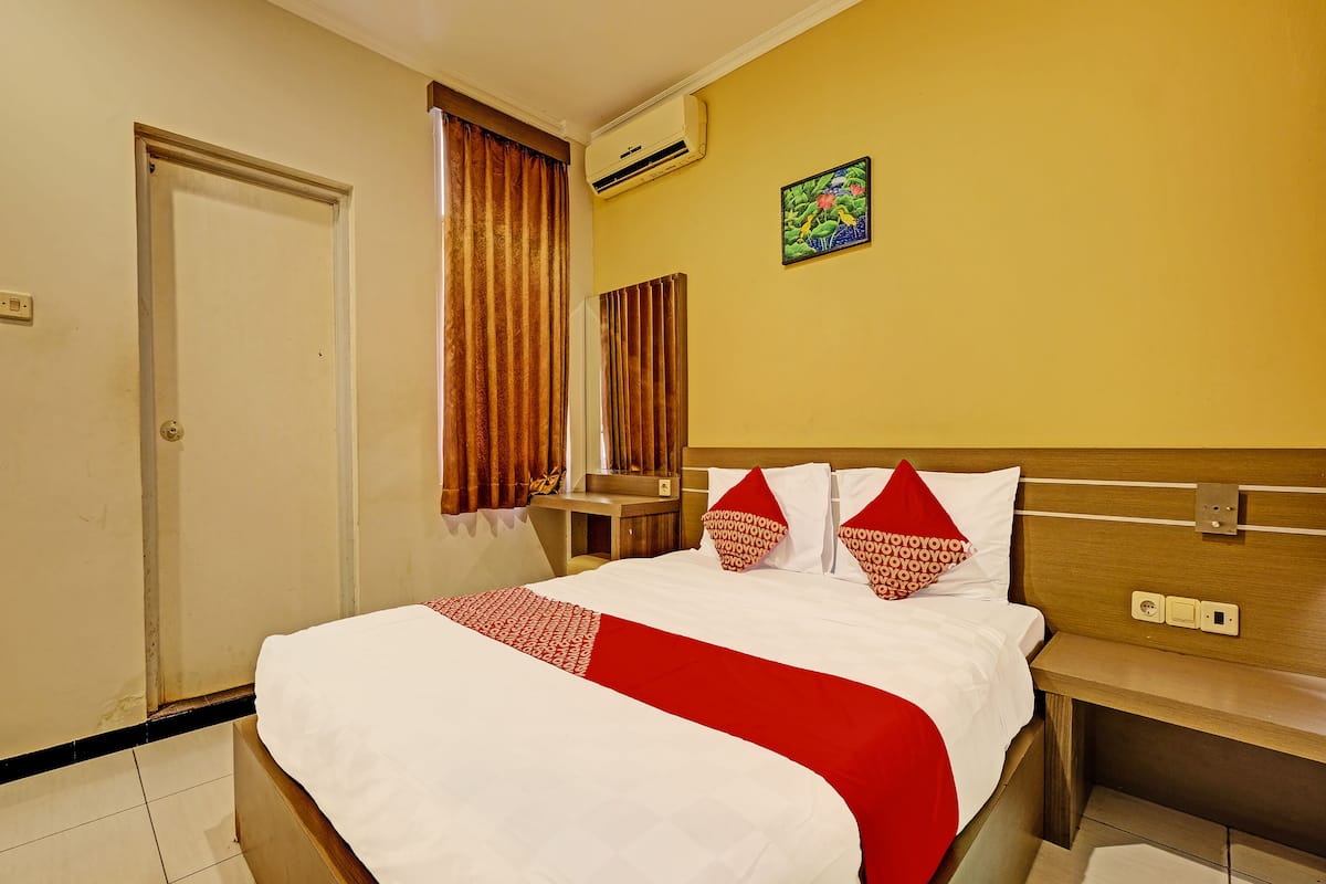 A hotel room with a bed, a bedside table, a mirror, an air conditioning unit, and a wall-mounted artwork.
