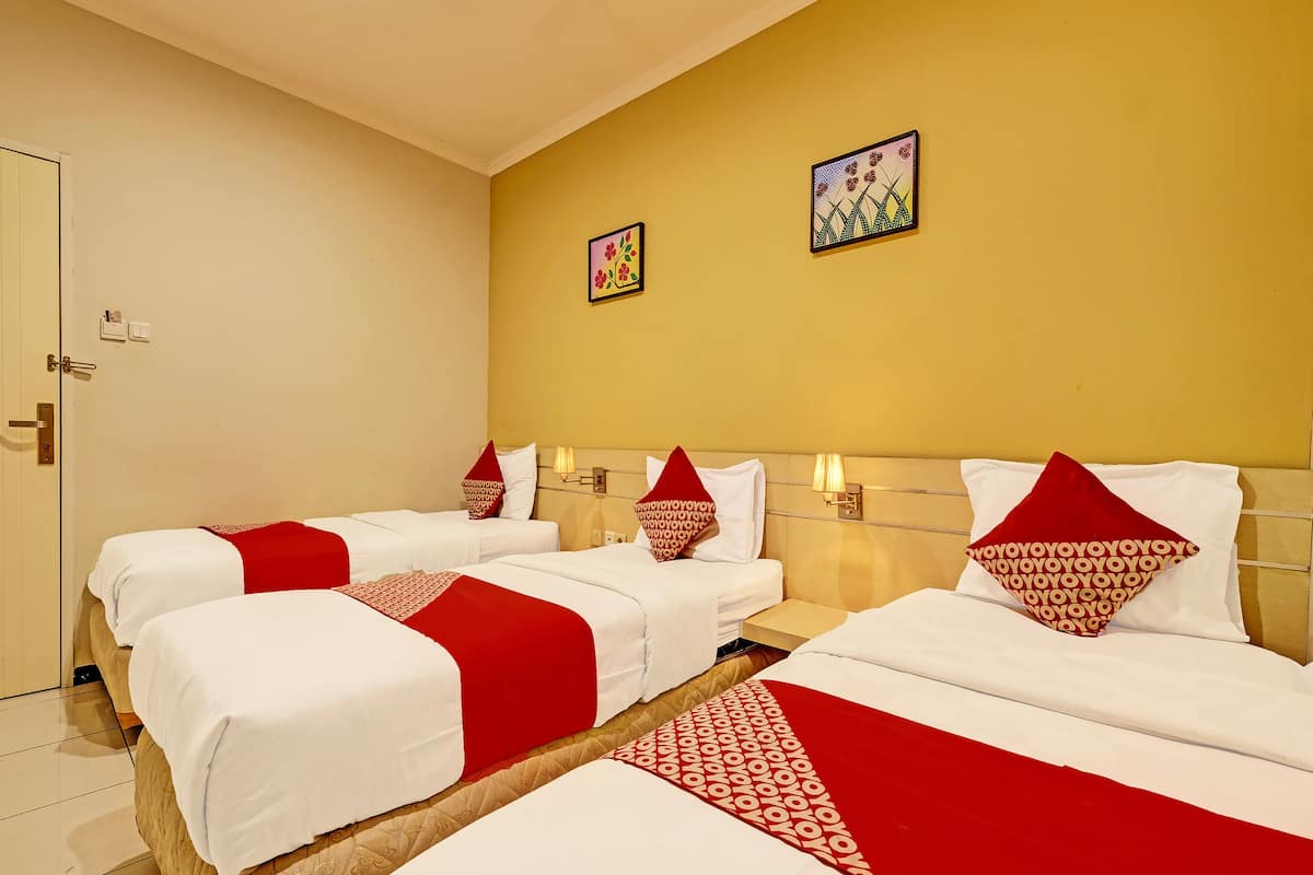 A hotel room with three beds, each with white bedding and red accents. There are two wall-mounted lamps and two framed artworks on the yellow walls.