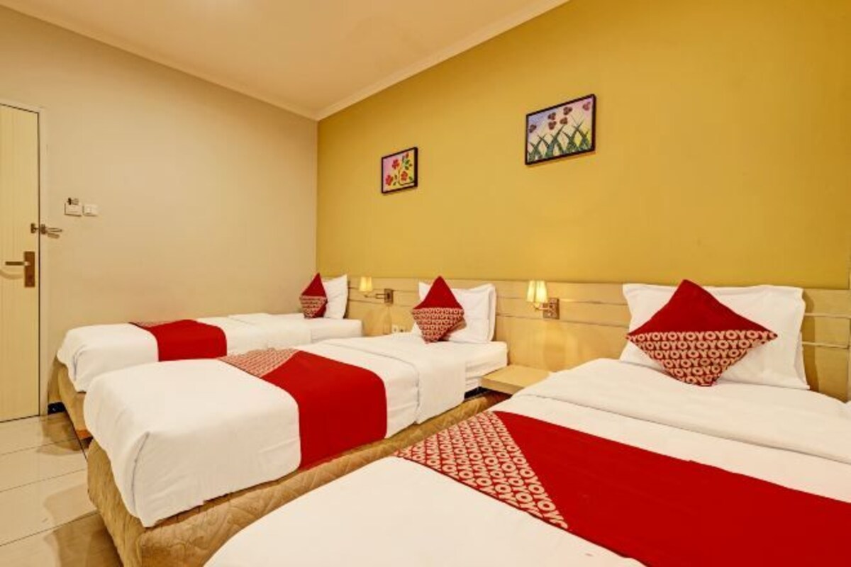 A hotel room with three beds, each with white bedding and red accents, a wooden headboard, and two wall-mounted artworks.