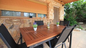 Pared exterior de piedra con una mesa y sillas de madera, una ventana con persianas y una puerta.