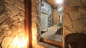 Interior de paredes de piedra con una escalera que lleva a una puerta, un sillón de mimbre y una lámpara fijada a la pared.