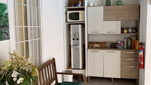 Een compacte keuken met een magnetron, koelkast en houten kastjes. Een eettafel met een groene kussen en bloemenboeket is zichtbaar.