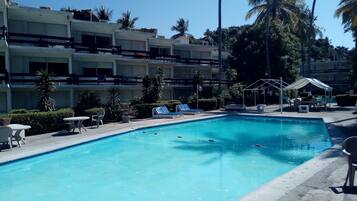 Una piscina con sillones de descanso y un área de estar techada, rodeada de palmeras y un edificio de varios pisos.