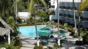 A resort pool area with lounge chairs and umbrellas, surrounded by palm trees and a building with multiple balconies. - Hotel La Jolla (Acapulco)