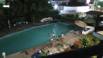 Una piscina con gente alrededor, rodeada de vegetación y edificios.
