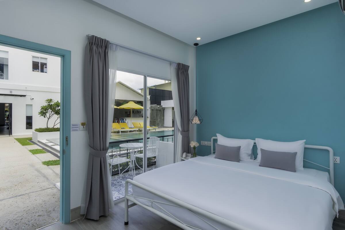 Ein modernes Hotelzimmer mit einem großen Bett, Blick auf den Poolbereich und einem Balkon mit Sitzgelegenheit.