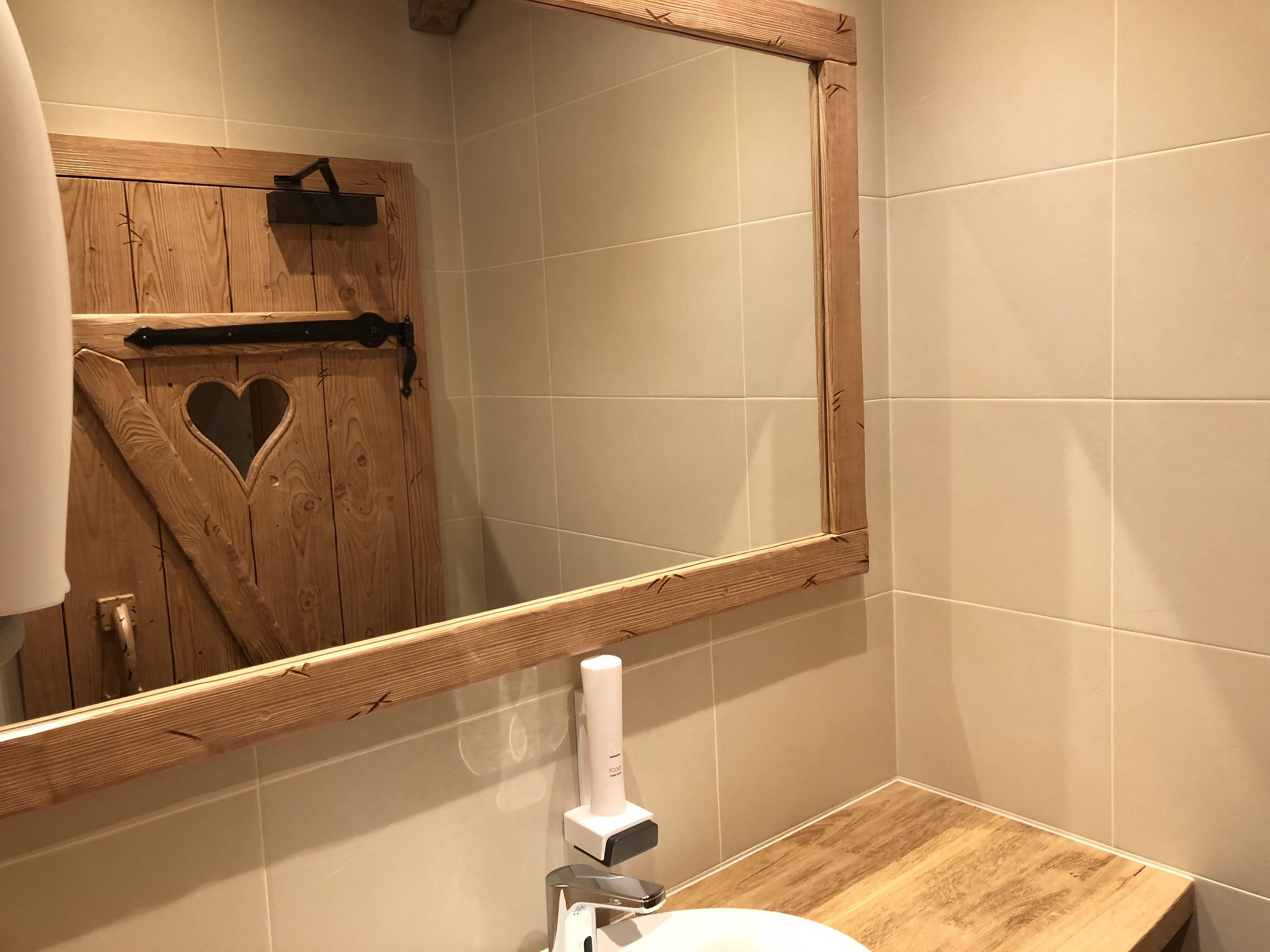 Een badkamer met een houten deur, een hartvormige uitsparing, een spiegel, een witte wastafel en een zeepdispenser.