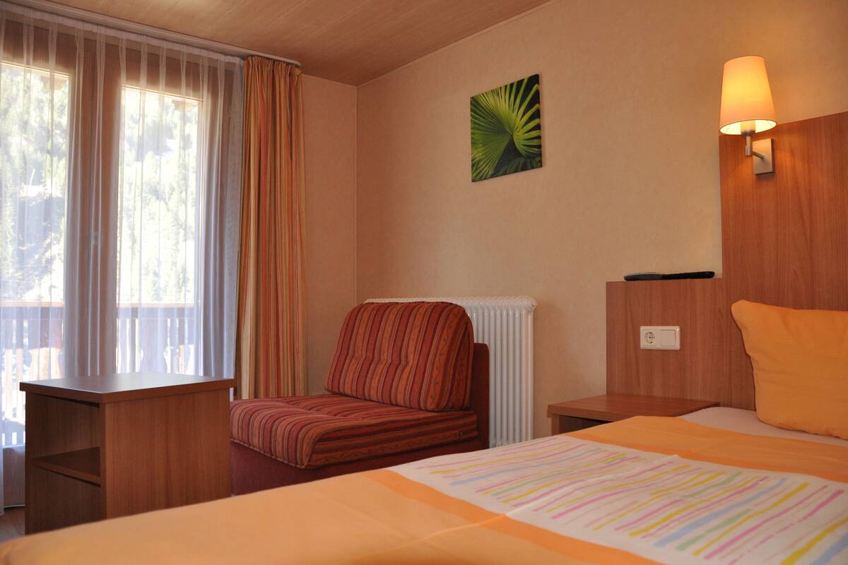 A hotel room with a bed, a chair, a desk, a radiator, and a wall-mounted lamp.