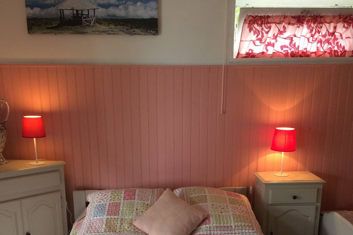 Ein Schlafzimmer mit gesteppter Bettdecke, zwei Nachttischen inklusive Lampen, einem Fenster mit Blumenmuster-Vorhang und einem Bild an der Wand.
