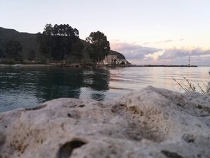 A coastal scene with a rocky foreground, calm water, and a distant view of a hill with trees and a structure. - Villa Marian (Apokoronas)