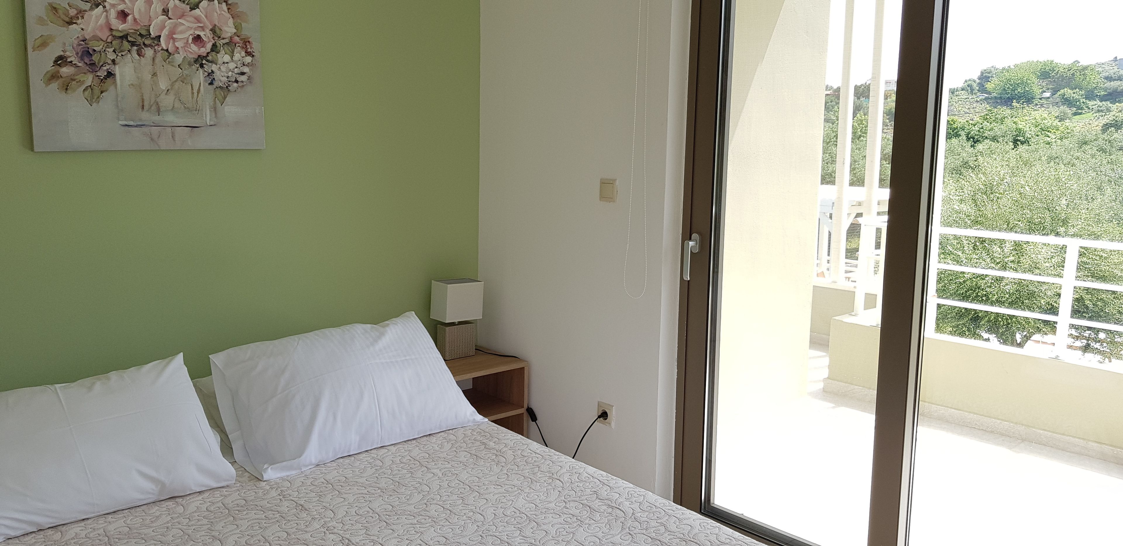 Una camera da letto con un letto, cuscini, un comodino con una lampada, un quadro al muro e un balcone con vista sul verde.