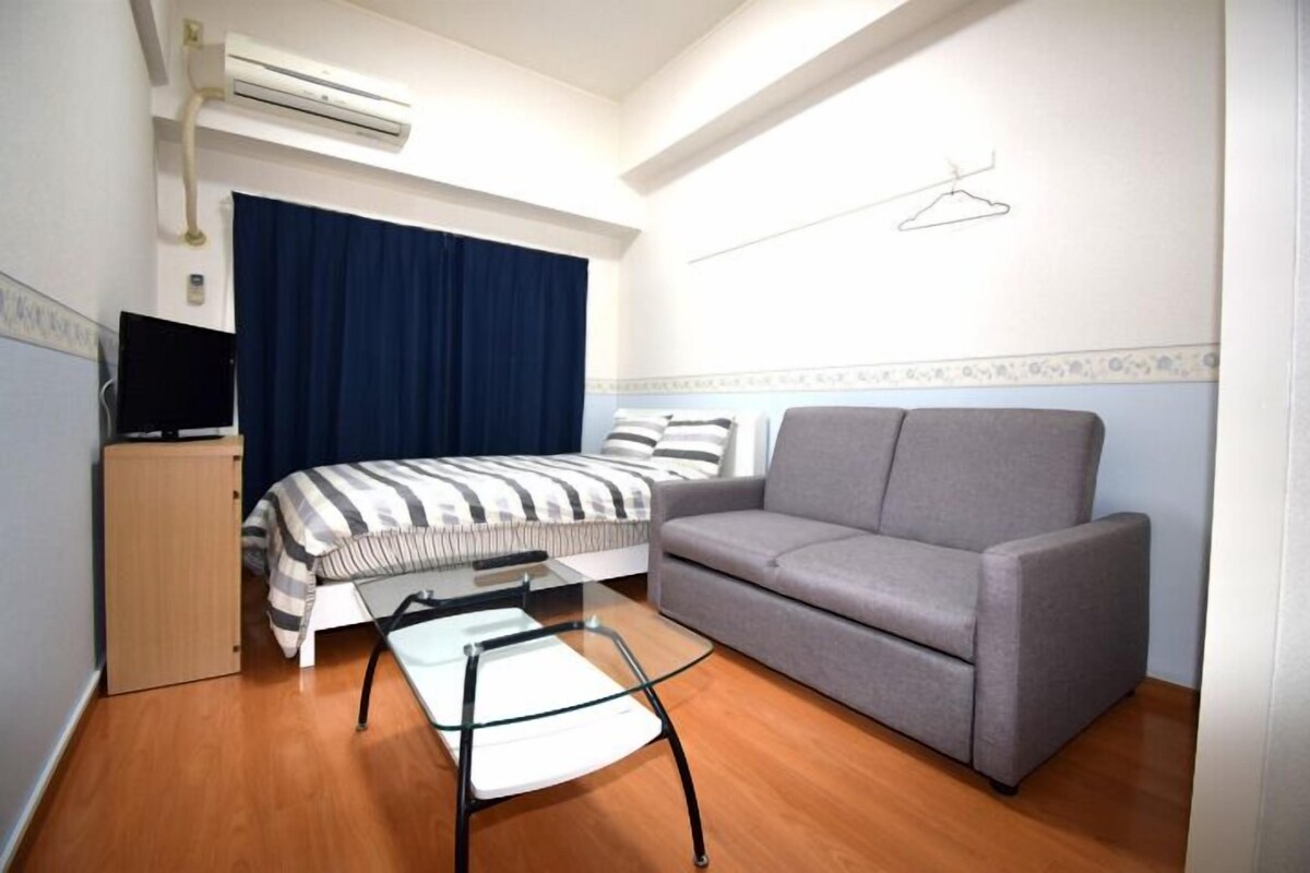 A hotel room with a bed, a grey sofa, a small table, a TV, and an air conditioning unit.