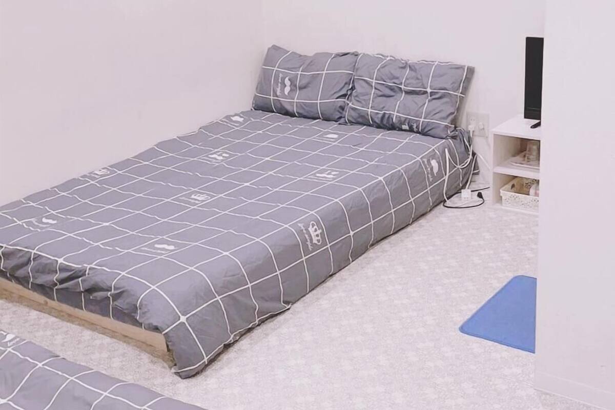 A small, single-bed room with a grey bedspread and matching pillows, a small desk with a computer, and a blue mat on the floor.