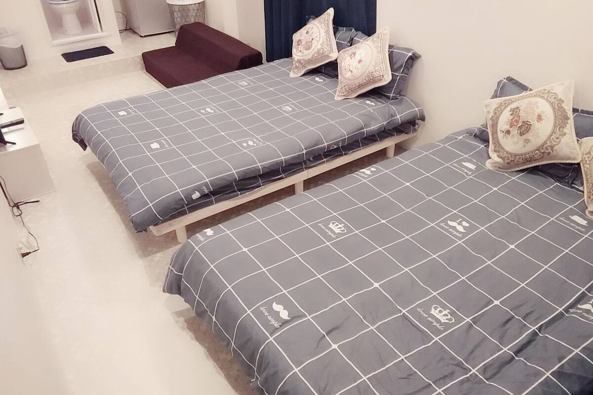 A bedroom with two beds, each with a grey checkered bedspread and matching pillows. There is a small wooden bench and a trash bin in the corner.