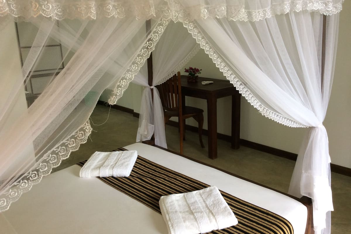 Quarto com cama, uma cadeira, uma mesinha e um dossel decorativo.