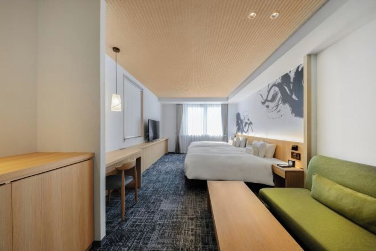 Ein Hotelzimmer mit einem Bett, einem grünen Sofa, einem Schreibtisch und einem großen Kunstwerk an der Wand.