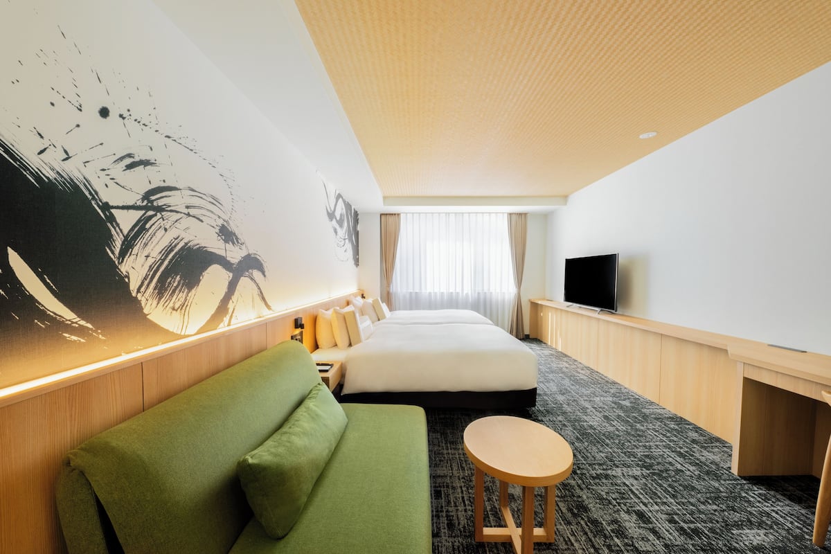 Een hotelkamer met een groot bed, een groene bank, een klein rond tafeltje, een flatscreen televisie en een groot abstract schilderij aan de muur.