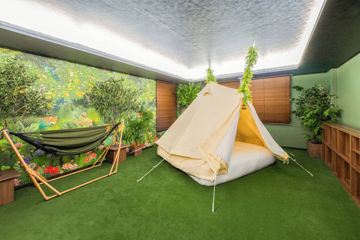 Een kamer met een vloer van kunstmatig groen gras, een hangmat, een tent en een muurschildering van een tuin.