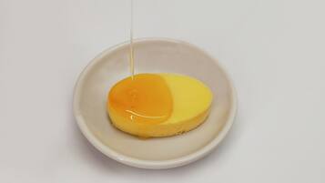 A single white bowl containing a yellow dessert with a glossy topping, being poured from a transparent container.