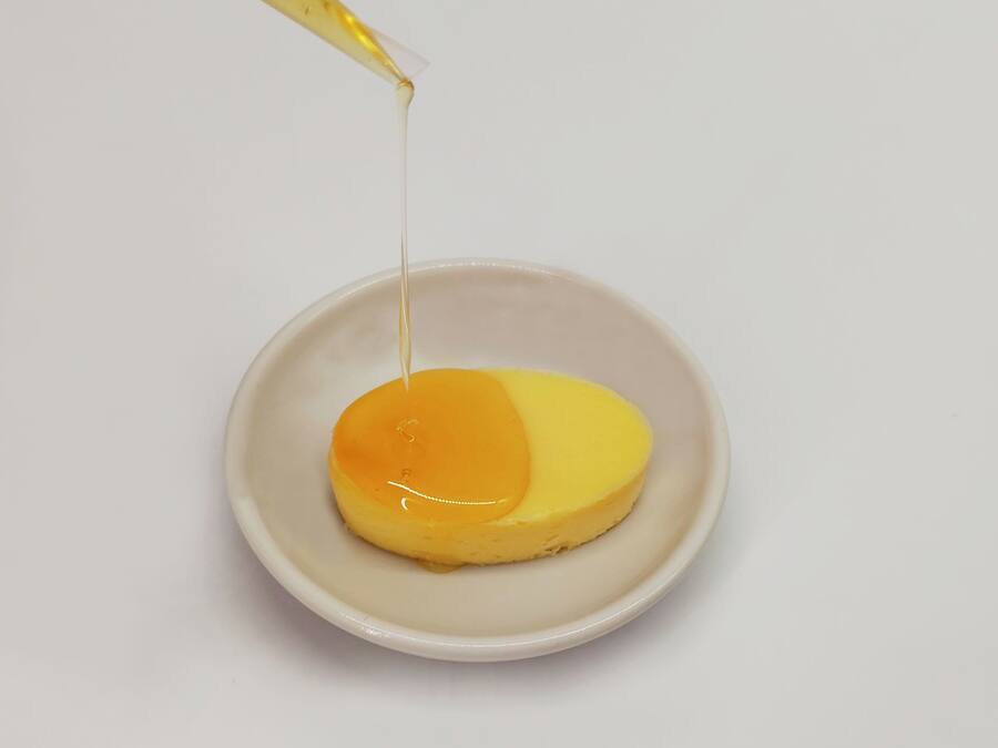 A single white bowl containing a yellow dessert with a glossy topping, being poured from a transparent container.