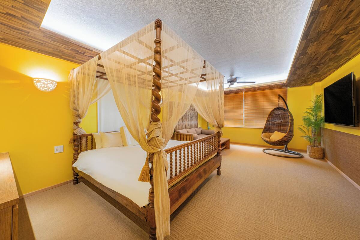 Een bed met hemelbed, een hangstoel, een televisie en een plafondventilator in een kamer met gele wanden en een houten plafond.