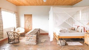 Een slaapkamer met een houten plafond, een bed met een witte hemel, een stenen vloer en een rieten stoel.