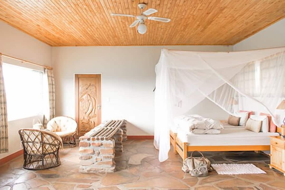 A bedroom with a wooden ceiling, a bed with a white canopy, a stone floor, and a wicker chair.