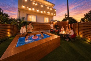 A swimming pool area with a wooden deck, surrounded by a fence, and a house with string lights in the background. - Grandioso Okinawa Villa Onna 2 (Onna)