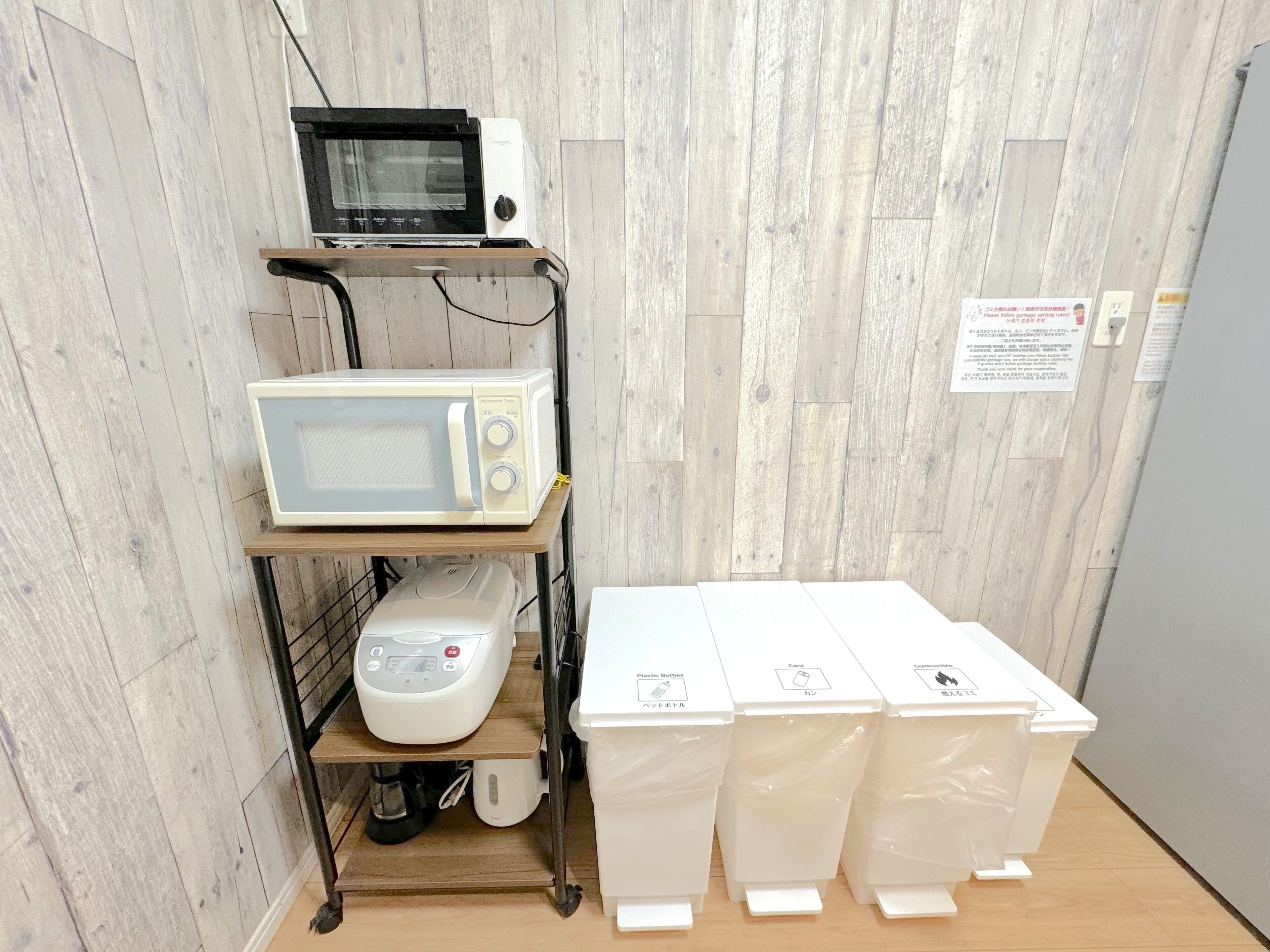 A kitchenette with a microwave, toaster, and coffee maker on a cart, and three trash bins labeled for different waste types.