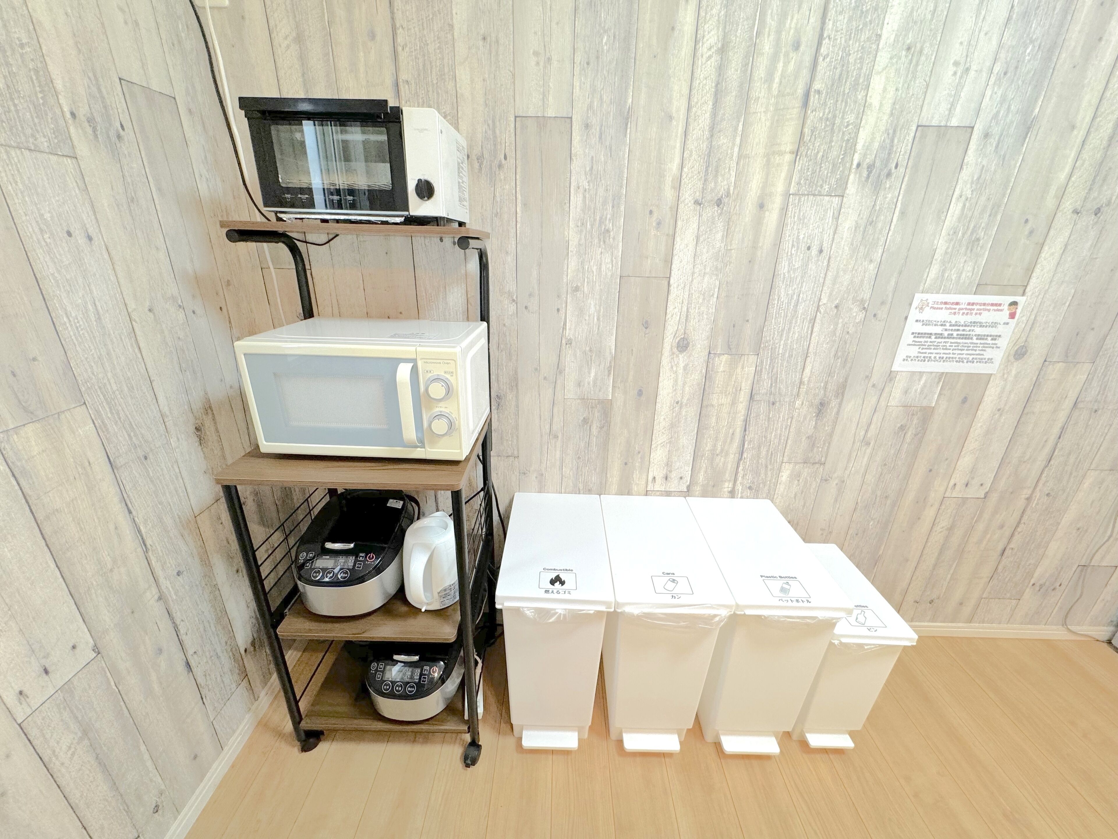 A kitchen area with a microwave on a shelf, a kettle, and several containers on the floor.