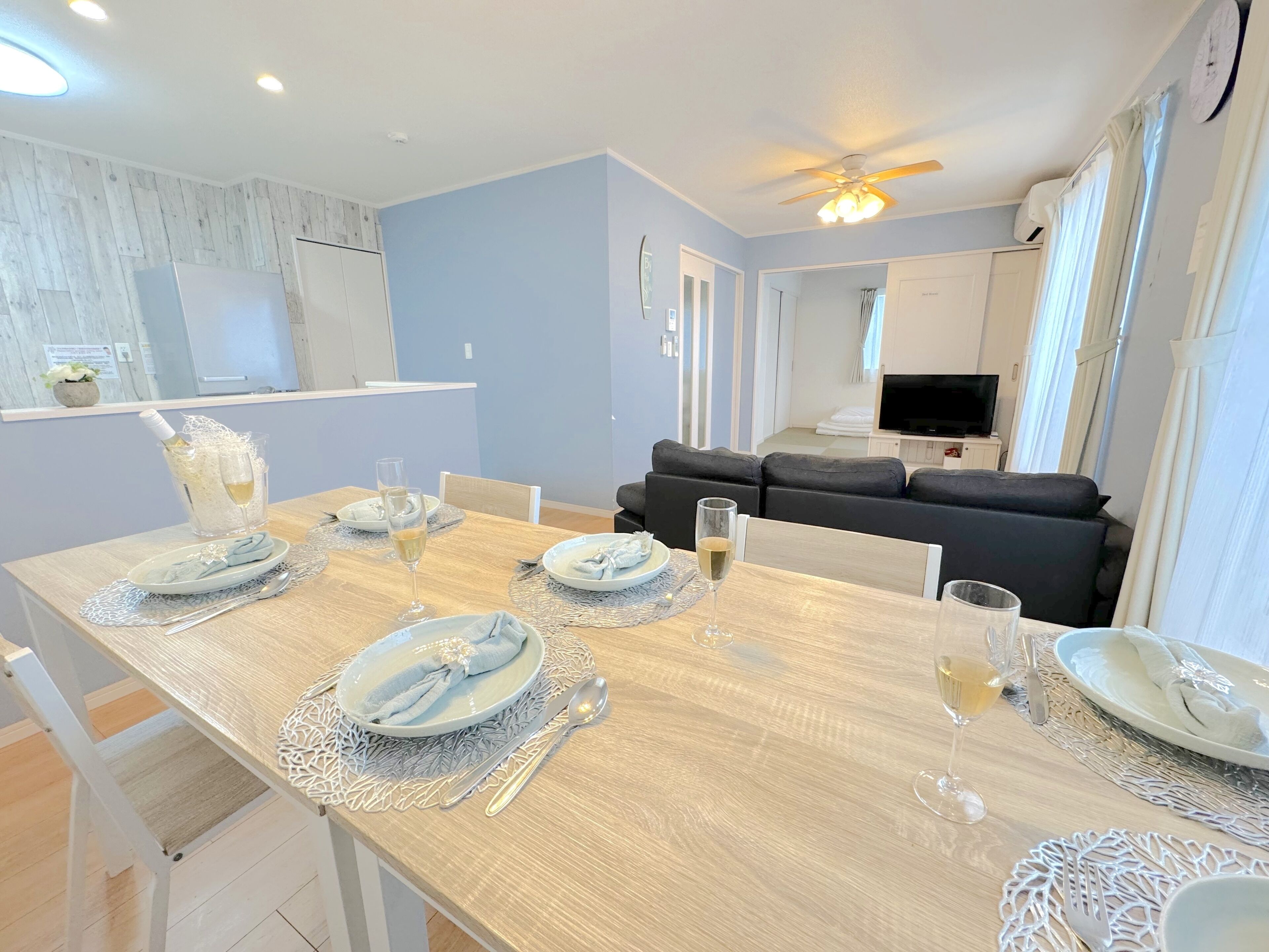 A dining area with a wooden table set for four, including plates, glasses, and cutlery. A ceiling fan and recessed lighting are visible above the table.