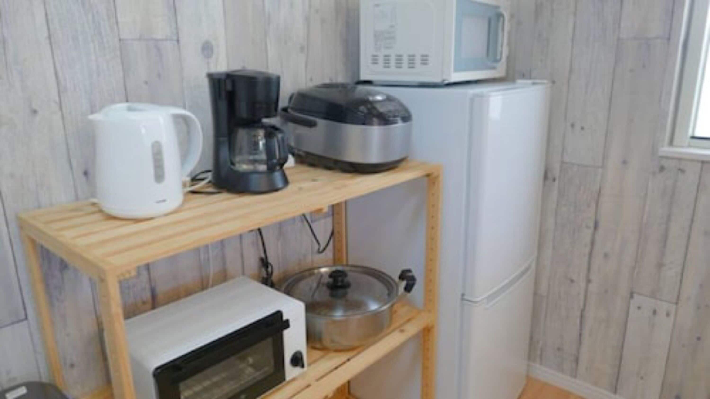 A kitchenette with a microwave, coffee maker, and a slow cooker on a wooden shelf, and a refrigerator in the background.