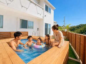 A swimming pool area with a wooden deck, a white building with windows, and a view of greenery in the background. - Grandioso Okinawa Villa Onna 2 (Onna)