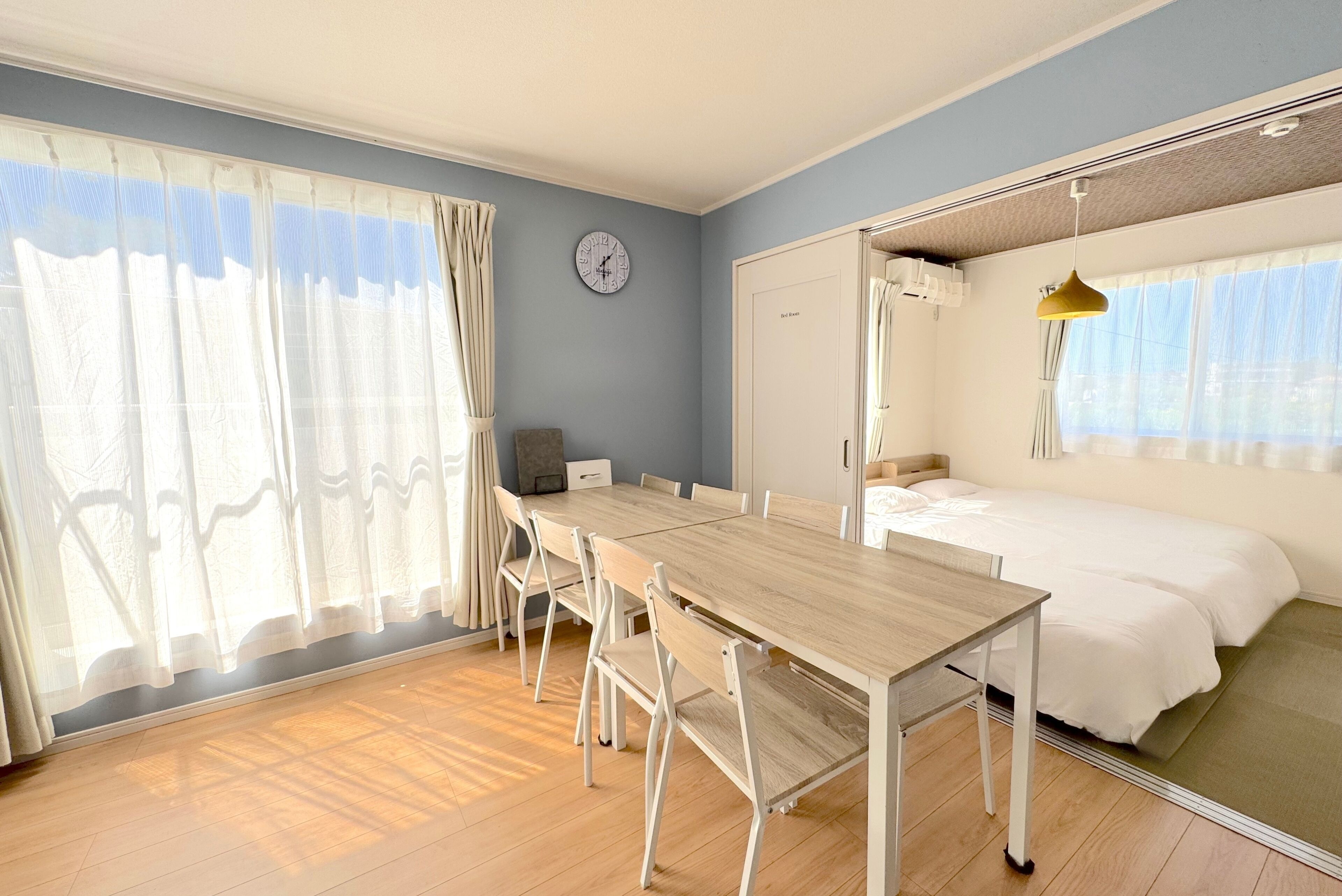 A room with a wooden dining table, chairs, a bed, a wardrobe, and a clock on the wall.