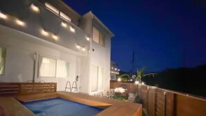 A swimming pool with a wooden deck, surrounded by a fence, and adjacent to a building with windows and outdoor lighting. - Grandioso Okinawa Villa Onna 2 (Onna)