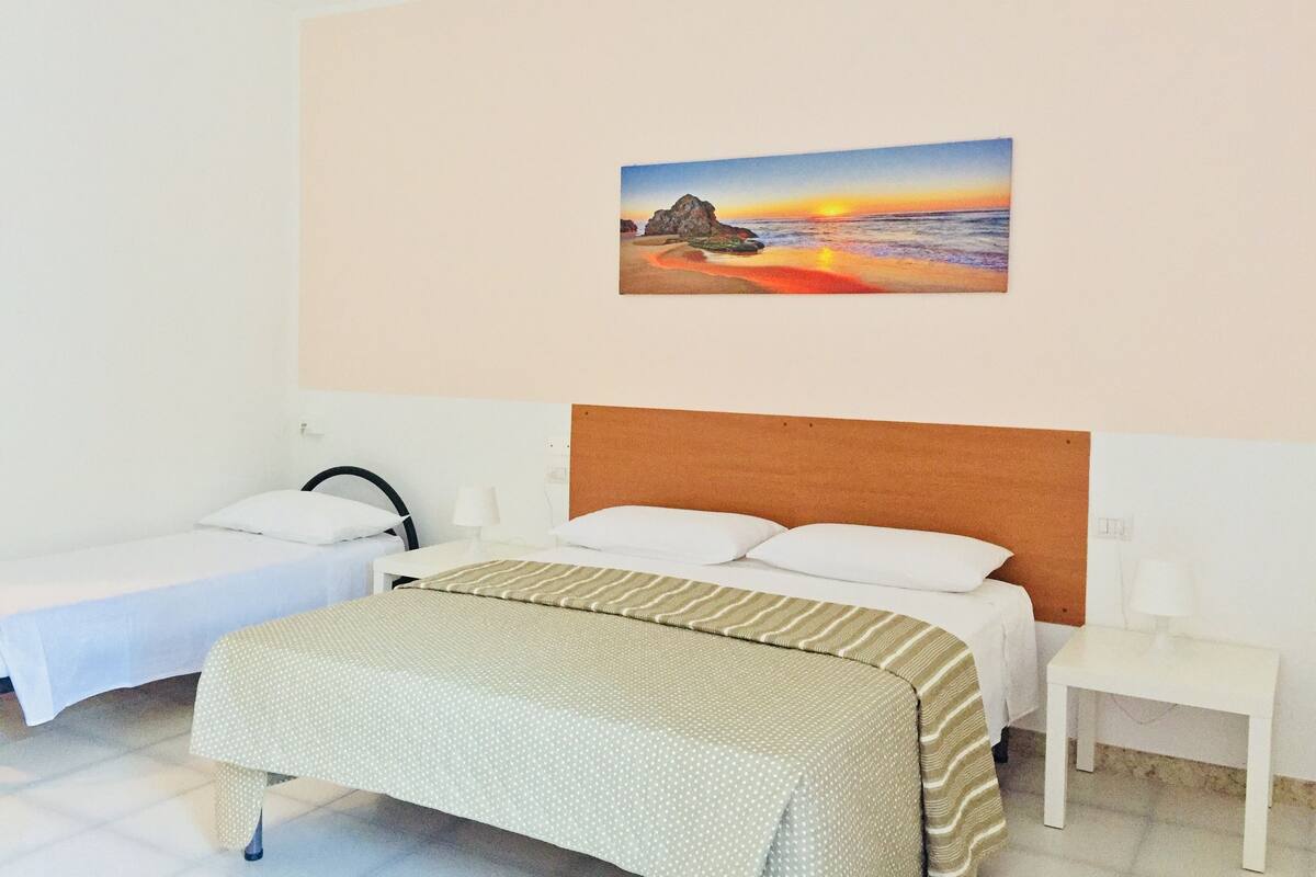 Habitació d'hotel amb un llit, un llit addicional o una llitera, una tauleta de nit i una pintura del sol ponent sobre l'oceà.