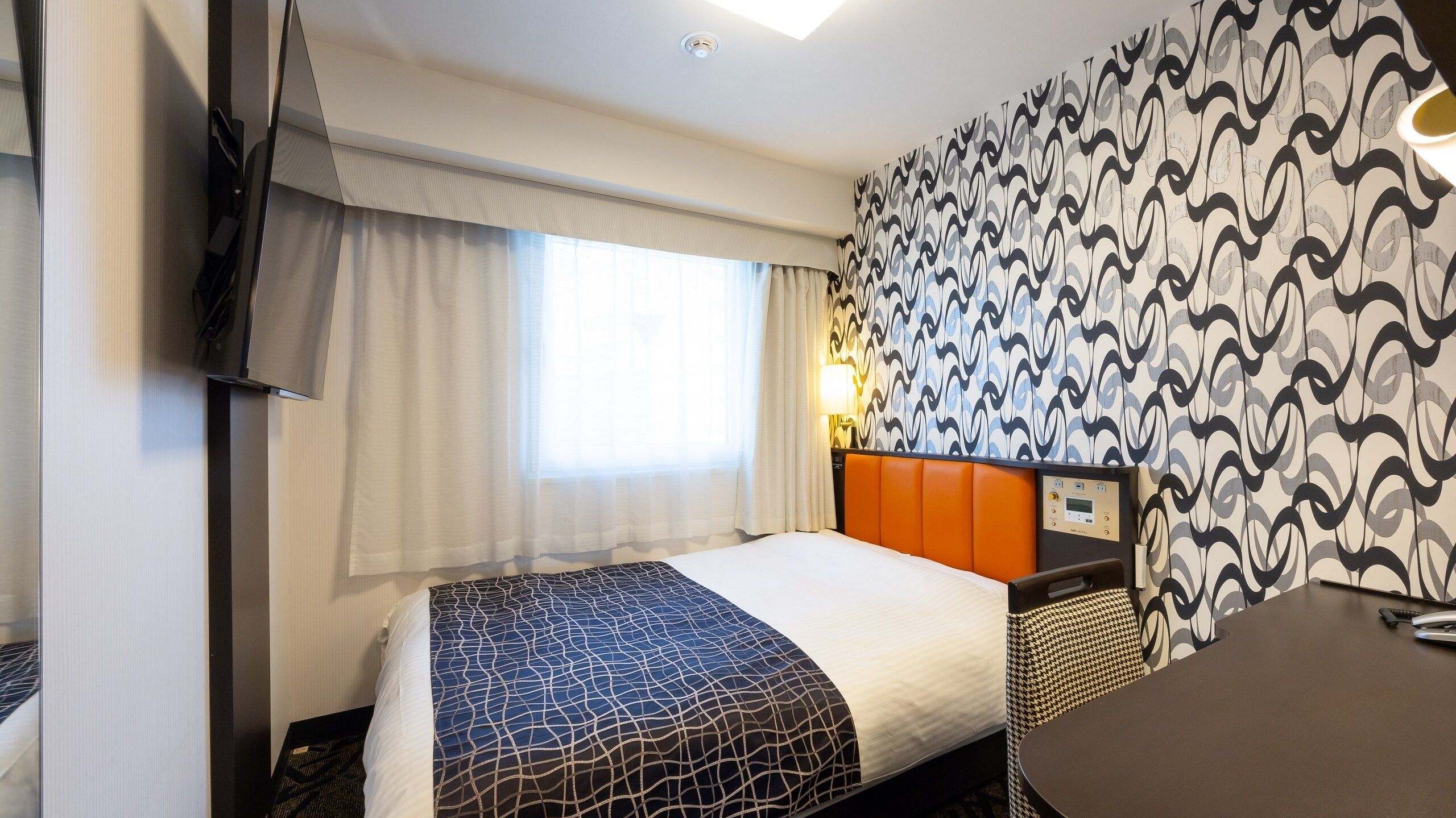 A hotel room with a bed, a desk, a TV, and a patterned wall.